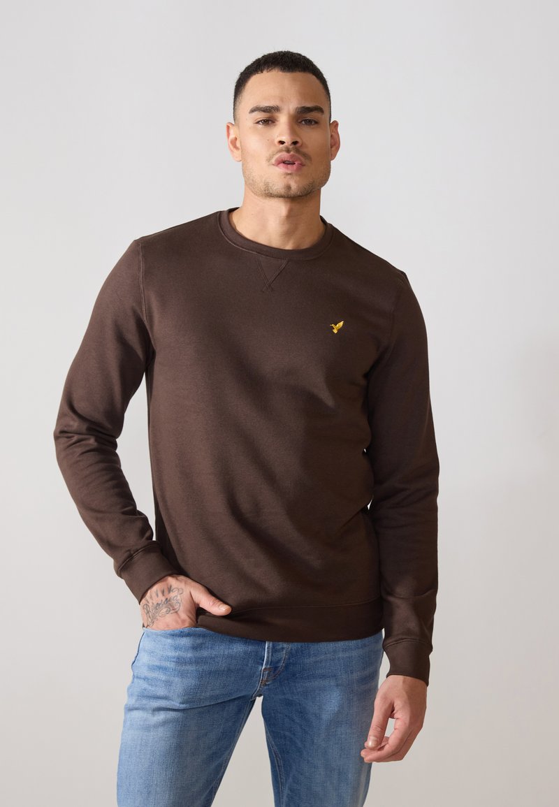 Brown cotton sweatshirt with long sleeves, crew neckline, and a small yellow logo on the chest, paired with light blue jeans.