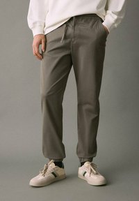 Person som bär grå joggingbyxor, vit sweatshirt och vita sneakers med gröna ränder, stående mot en enfärgad grå bakgrund.