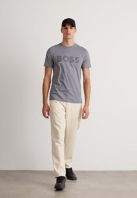 Hombre caminando, vistiendo una camiseta gris de "BOSS", pantalones crema, zapatillas negras y una gorra negra, de pie contra un fondo blanco liso.
