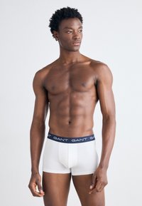 Boxer shorts blancos con una cinturilla azul marino que presenta el nombre de la marca "GANT" repetido. La tela es ajustada y de textura suave.