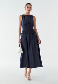 Abito maxi blu marine con vita arricciata, corpetto plissettato, dettagli volants e gonna fluida. Abbinato a una borsa bianca e sandali con cinturino.