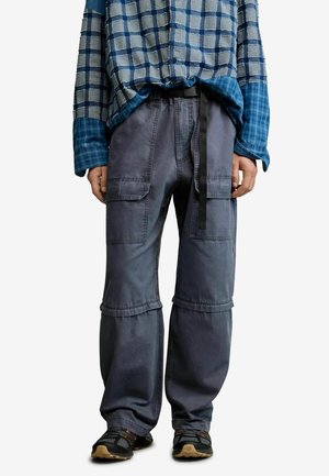 Persona con camisa azul a cuadros, pantalones cargo sueltos de color gris oscuro con múltiples bolsillos, cinturón negro y zapatillas negras de trail running.