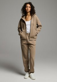 Beżowa hoodie z troczkiem i przednimi kieszeniami, w zestawie z dopasowanymi dresami zwężającymi się ku dołowi. Ubranie charakteryzuje się miękką teksturą i białymi sneakersami.