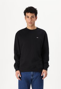 Sweatshirt noir en coton avec col rond, épaules descendues et poignets côtelés. Présente un petit logo coloré sur la poitrine. Associé à un jean bleu.