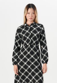 Femme aux cheveux raides portant une robe à manches longues noire et blanche à motifs avec un col, se tenant devant un fond blanc uni.