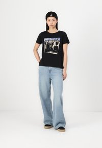T-shirt noir à manches courtes présentant un graphique imprimé d'un groupe de musique. Associé à un jean large bleu clair et des chaussures noires à enfiler.