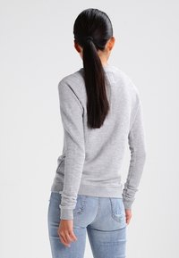 Donna con una lunga coda di cavallo scura, che indossa un maglione grigio chiaro a maniche lunghe e jeans in denim blu chiaro, in piedi di spalle.