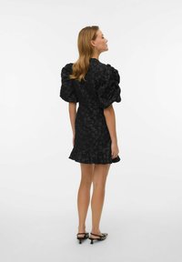 Črna obleka z nabranimi rokavi, cvetličnim vzorcem in nagubanim robom. Združena s črno-belimi čeveljci z ostrimi konicami, prikazanimi od zadaj.