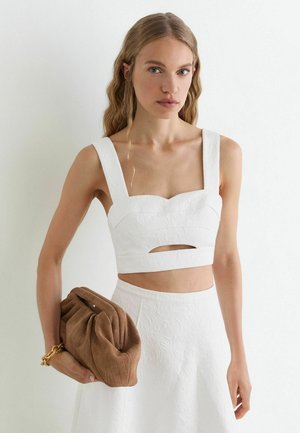 Žena s dlhými vlnitými blond vlasmi nosí biely textúrovaný crop top a sukňu, drží hnedú prešívanú kabelku so zlatým retiazkovým náramkom.