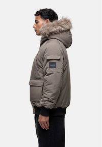 Giubbotto imbottito verde oliva con cappuccio in pelliccia sintetica, zip frontale e tasche laterali, polsini a costine e patch del logo sulla manica.