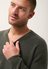 Olijfgroene V-halstrui, gemaakt van textuurmateriaal, met lange mouwen en ribgebreide manchetten. Hand voorzichtig geplaatst op de kraag.