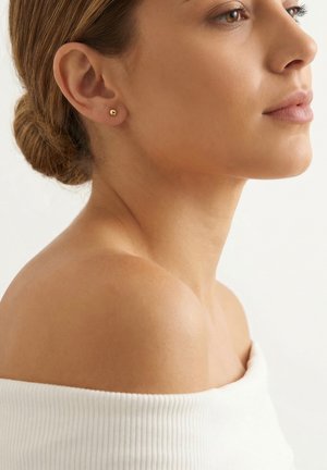 Jeune femme aux cheveux châtain clair en chignon, portant de petites boucles d’oreilles en or et un haut blanc sans épaules, tournée vers la droite sur un fond uni.