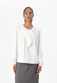 Blusa bianca con una scollatura smockata testurizzata e un fiocco al colletto. Maniche lunghe e ampie. Indossata con una gonna grigia a righe.