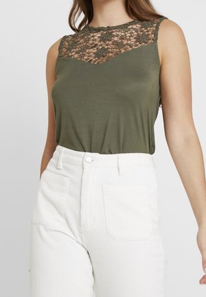 Vrouw draagt een mouwloze olijfgroene top met een bloemige kanten inzet en witte, hooggetailleerde corduroy broek.
