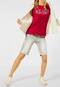 Röd ärmlös t-shirt med texten "READY set glow", kombinerad med ljusa denimshorts och vita sneakers. Öppen vit jacka synlig.