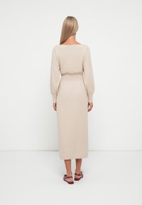 Abito midi in maglia beige con maniche a sbuffo lunghe, vita aderente e gonna dritta. Abbinato a sandali rossi. Texture liscia, design minimale.