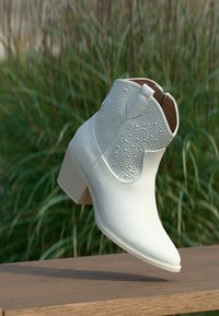 Λευκό δερμάτινο μποτάκι αστραγάλου με ανάγλυφη επιφάνεια, ταμπελάκι έλξης και στιβαρό τακούνι, τοποθετημένο πάνω σε ξύλινη επιφάνεια με πράσινο χλοοτάπητα στο βάθος.