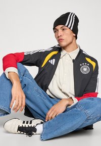 Svart jakke med fargerike aksenter (rødt, gult) og hvite striper, kombinert med en beige skjorte og dongeribukser. Svart lue og hvite joggesko.