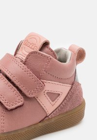 Růžová kožená bota s háčky na suchý zip, texturovaným nubukovým podpatkem a gumovou podrážkou. Na boku má trojúhelníkové výřezy.