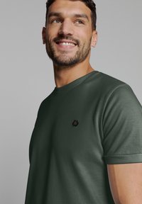 Donkergroen t-shirt met korte mouwen van soepel materiaal, met een zwart hexagonaal logo op de borst en een ronde hals.