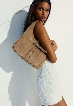 Young woman wearing a white textured dress carrying a large tan suede shoulder bag with front pockets and a buckle strap.