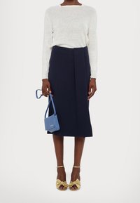 Jupe portefeuille marine, pull en tricot léger blanc, sandales à talons dorées avec un détail nœud, et un petit sac à main bleu.