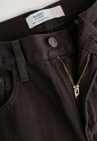 Dark brown denim pants with a partially open brass zipper, metal button, belt loop, and a white label reading "BAGGY MID RISE MADE IN BANGLADESH."