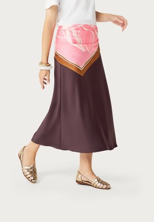 Mujer caminando con falda midi marrón, pañuelo rosa estampado atado a la cintura, blusa blanca, sandalias planas doradas y pulseras doradas y blancas.