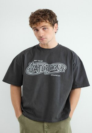 Mladý muž s kučeravými vlasmi má na sebe čierne oversized tričko s nápisom "Get your Saturday groove on", ruky v vreckách olivovo zelených nohavíc.