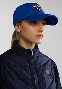 Cappello blu con una patch rettangolare con la bandiera norvegese, abbinato a un giubbotto imbottito blu navy con chiusura a zip e dettaglio del logo rotondo.