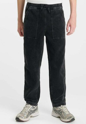 Schwarze Cordhose mit Vordertaschen und Kordelzugbund, getragen von einer stehenden Person mit weißen und grauen Laufschuhen.