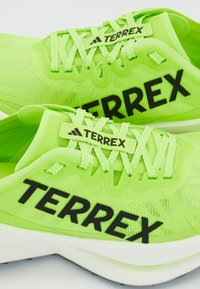 Zelene sportske tenisice s teksturiranim mrežastim gornjištima, crnim oznakama "TERREX" i bijelim potplatima. Imaju sjajne vezice i aerodinamičan dizajn.
