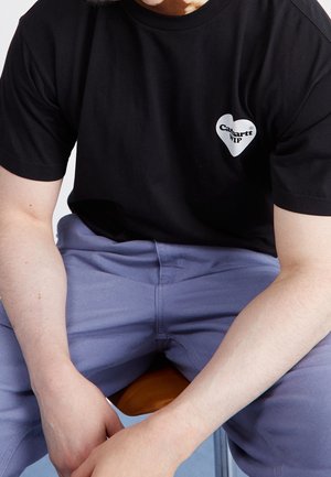 Personne assise portant un T-shirt noir avec un logo "Carhartt WIP" en forme de cœur blanc et un pantalon bleu clair, les mains reposant entre les jambes.