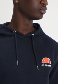 Sudadera azul marino de algodón, con capucha ajustable, herrajes plateados y un logo de ellesse en rojo y blanco en el lado izquierdo del pecho.