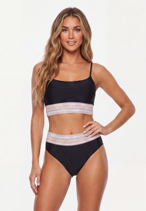 Fiatal nő hosszú hullámos hajjal, fekete bikiniben mintás szegéllyel, egyik kezét csípőjén tartva, egyszínű háttér előtt áll.