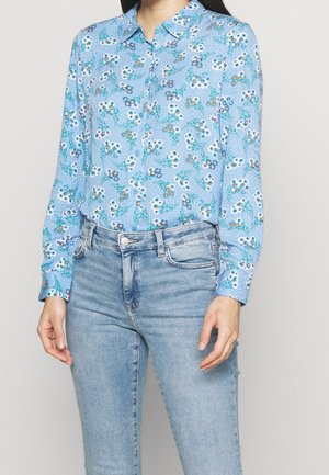 Lichtblauwe button-up shirt met een bloemenpatroon in wit en oranje, gecombineerd met lichtdenim jeans. Zachte stof, lange mouwen en kraagdetails.