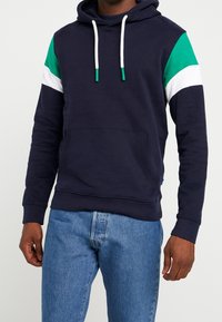 Felpa blu navy con accenti geometrici verdi e bianchi sulle spalline, cappuccio con lacci e tasca a marsupio. Tessuto morbido con polsini a costine.