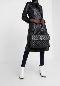 Černý kožený trenčkot s dvouřadým zapínáním, doplněný černou prošívanou kabelkou s řetízkovým popruhem a dekorativními kovovými detaily.