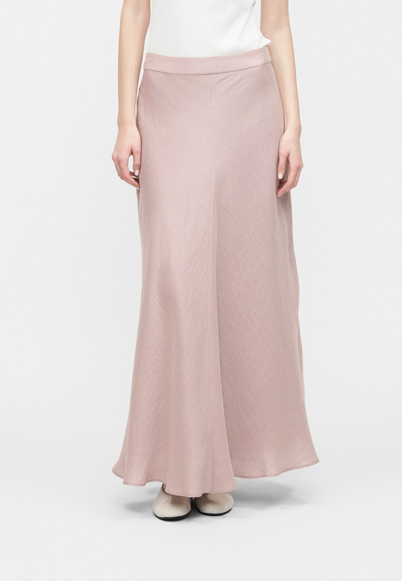 Γυναίκα που φοράει μια μακριά φούστα σε ανοιχτό ροζ χρώμα με ψηλή μέση, συνδυασμένη με λευκά παπούτσια και αμάνικο λευκό τοπ, στέκεται με τα χέρια χαλαρά.