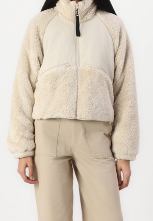 Mujer vistiendo una chaqueta de forro polar beige con cremallera frontal y pantalones beige a juego, de pie frente a un fondo blanco liso.