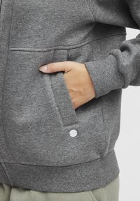 Gray zip-up sweatshirt with a textured finish, featuring a front pocket and a round metal accent on the hem.