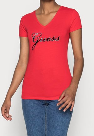 Woman wearing a red V-neck T-shirt with "Guess" in rhinestone lettering and blue jeans, standing against a plain background.