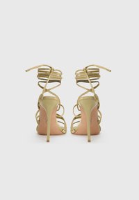 Sandales à talons hauts avec lanières en or métallisé. Présente des fines lanières avec un design enroulé et un talon aiguille élancé. Design à dos ouvert.