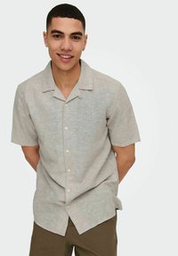 Camisa de manga corta de lino gris claro con un diseño de botones, que presenta un cuello y una textura sutil, llevada sobre un pantalón beige.