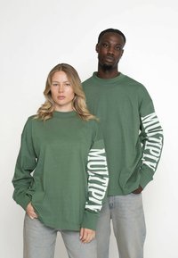 Grønne sweatshirts med lange ærmer har en høj halsudskæring og hvid tekst "MULTIPLY" langs ærmerne, parret med lyse denimjeans.