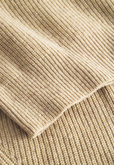 Bežani pleteni materijal s teksturiranim rebrastim uzorkom, koji ima mekan i topao izgled te suptilne varijacije boje u cijelom svom opsegu.