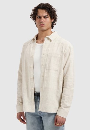 Man draagt een gestructureerd beige overhemd met knopen over een wit T-shirt, lichtblauwe jeans en gouden ringen aan zijn vingers, staand tegen een effen achtergrond.