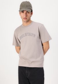 Γκρι βαμβακερό T-shirt με καμπυλωτό λογότυπο "BELSTAFF", συνδυασμένο με μια φθαρμένη πράσινη καπέλο που έχει το κείμενο "Won" κεντημένο.