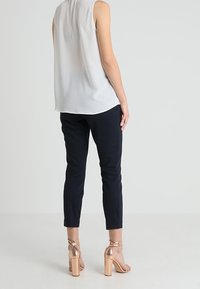 Top senza maniche di un grigio chiaro, pantaloni blu fitted e sandali con tacco a blocco color sabbia con cinturino alla caviglia, che mostrano una silhouette moderna e slanciata.