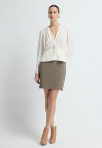 Cremfarbene Bluse mit tiefem V-Ausschnitt und drei goldenen Knöpfen an der Taille, kombiniert mit einem strukturierten beigen Rock und hellbraunen Stiefeletten.